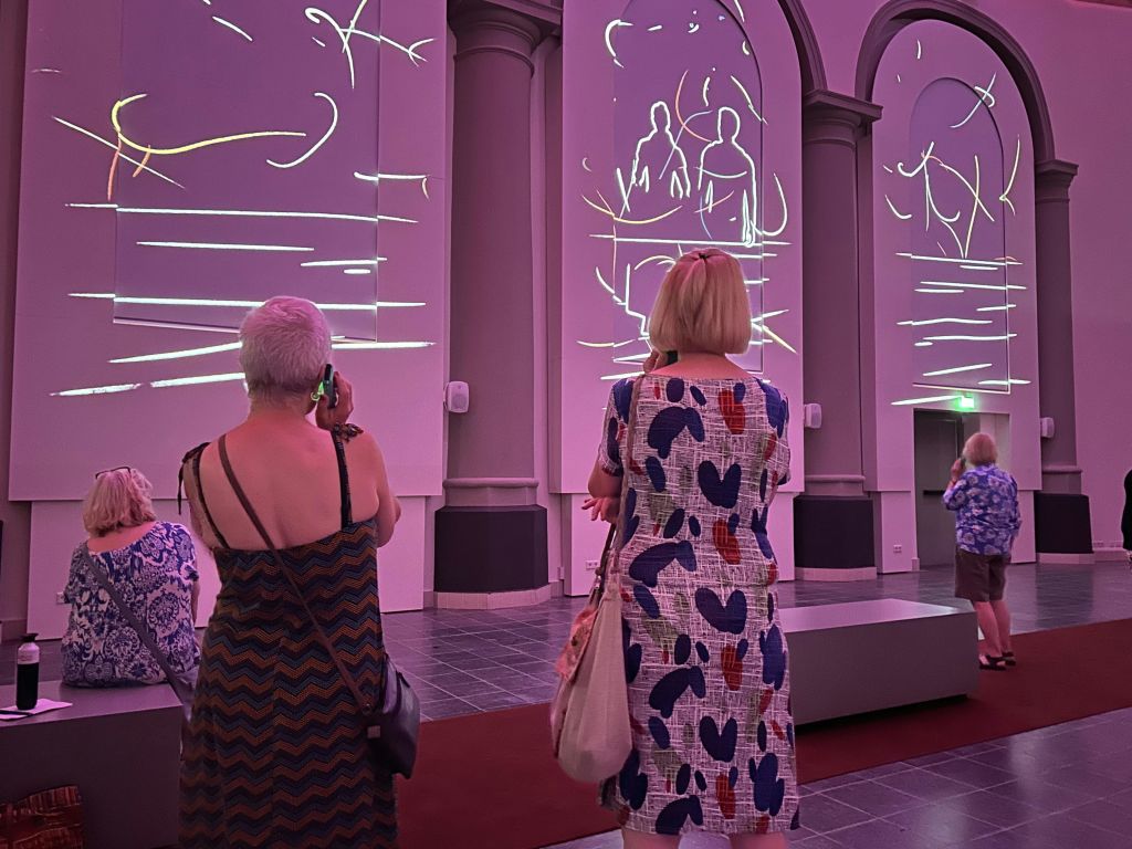 보라색 조명이 있는 갤러리의 사람들은 대형 벽 패널에 투영된 추상적인 흰색 선 예술을 감상합니다.