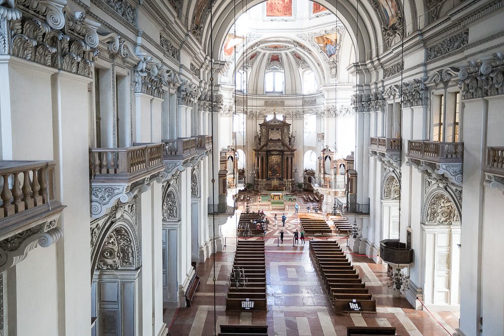 화려한 건축물, 높은 아치형 천장, 목조 퓨, 제단 근처를 걷는 몇 명의 방문객이 있는 넓은 대성당.