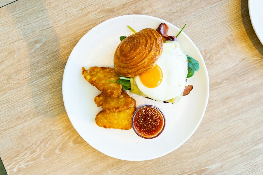 크루아상, 햇살 가득한 달걀, 베이컨, 해시 브라운, 그린, 작은 디핑 소스 한 그릇이 담긴 아침 식사용 접시입니다.