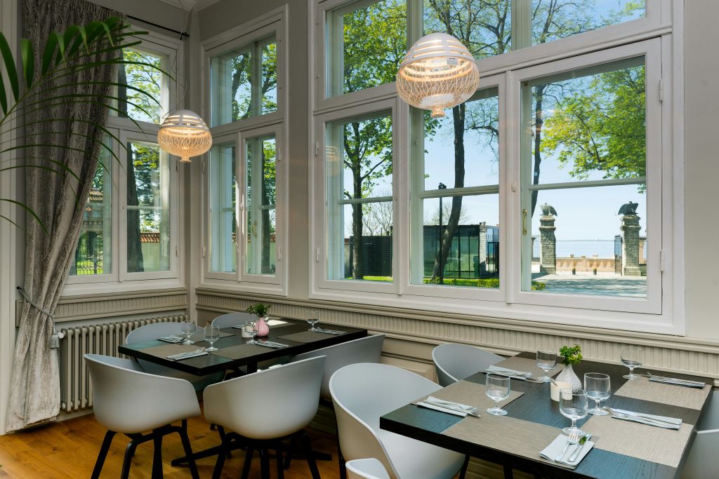 창문이 있는 햇살이 내리쬐는 식사 공간, 흰색 의자, 유리잔과 식기류가 놓인 어두운 테이블, 걸이식 램프, 바깥에 보이는 나무.