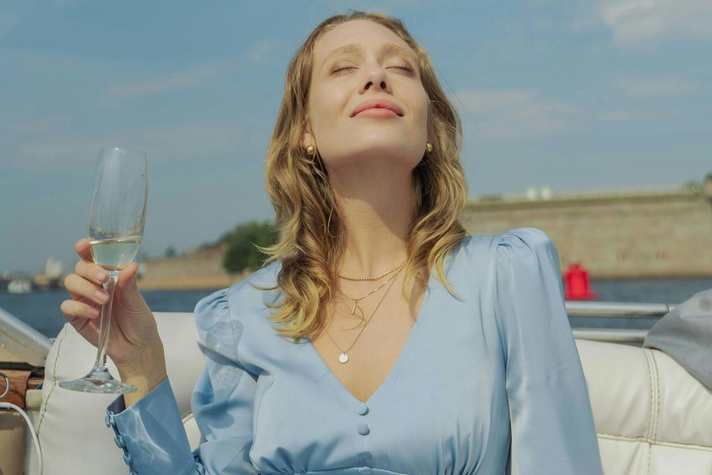 밝은 파란색 드레스를 입은 여성이 와인잔을 들고 눈을 감은 채 태양을 즐기고 있습니다. 배경은 부분적인 야외 장면을 보여줍니다.