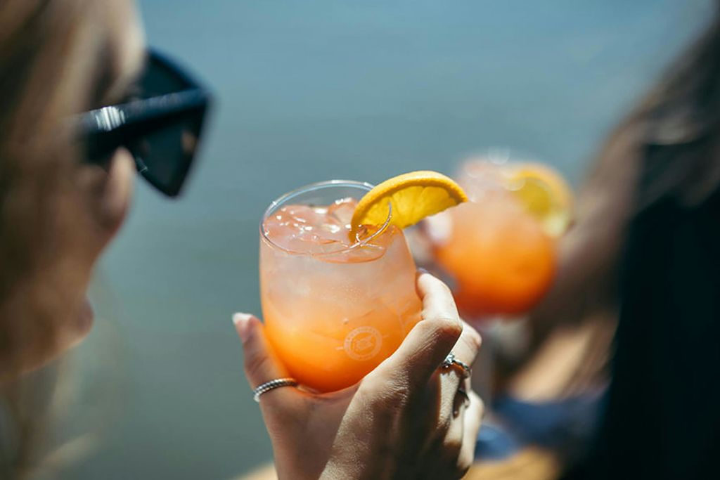 레몬 장식과 함께 오렌지 칵테일 한 잔을 들고 있는 사람이 배경에 또 다른 유사한 음료가 보입니다.