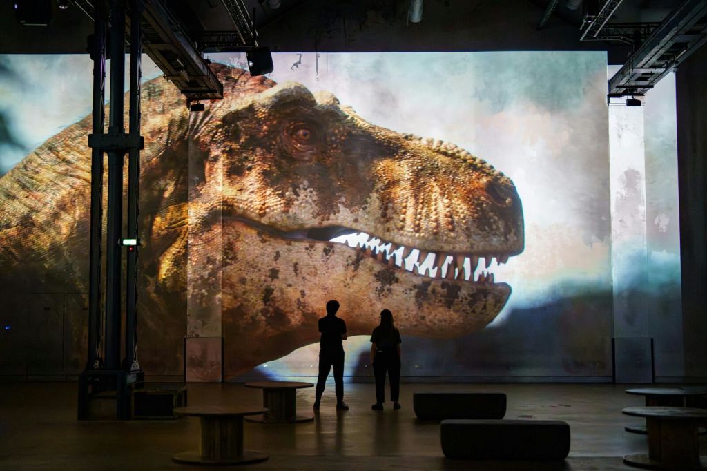 두 사람이 어두운 방에 서서 화면에 공룡의 머리가 커다랗게 투영된 것을 관찰하고 있습니다.