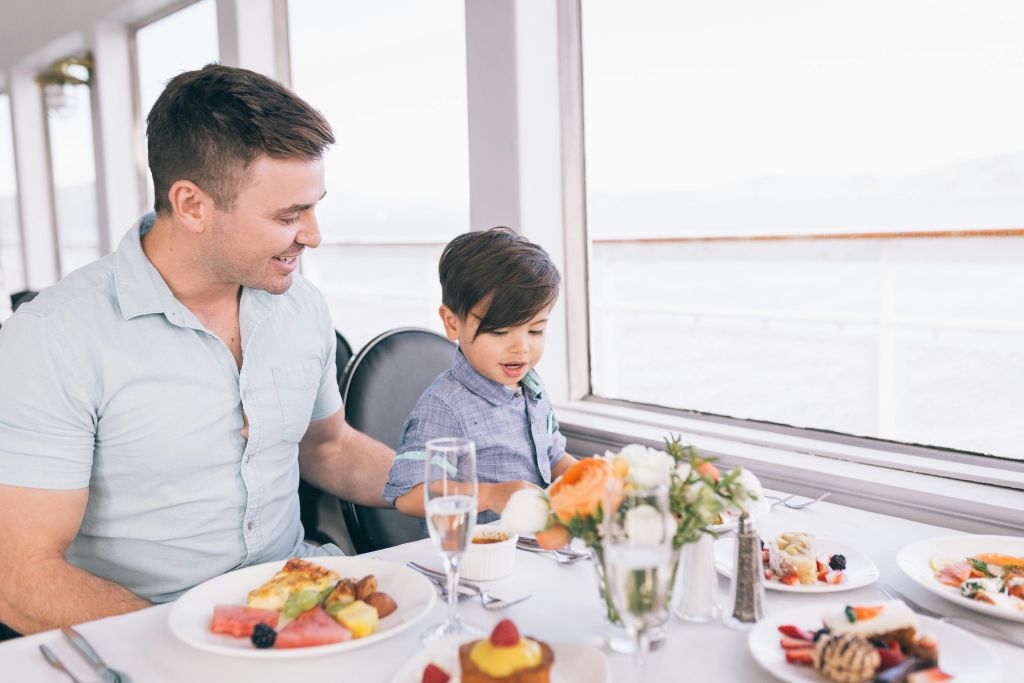 한 남자와 어린 소년이 창가에서 음식과 꽃 접시가 놓인 테이블에 앉아 서로 웃으며 교감하고 있습니다.