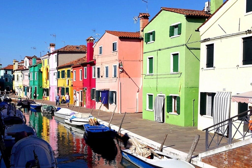 운하를 따라 밝은 색상의 건물과 배가 정박해 있고 맑고 푸른 하늘 아래 보도를 걷는 사람들이 몇 명 있습니다.