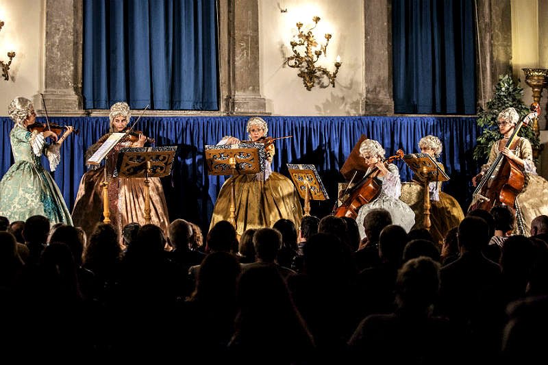 마침표 의상을 입은 네 명의 뮤지션이 관객 앞에서 무대에서 현악기를 들고 공연합니다. 파란색 커튼과 어두운 조명.