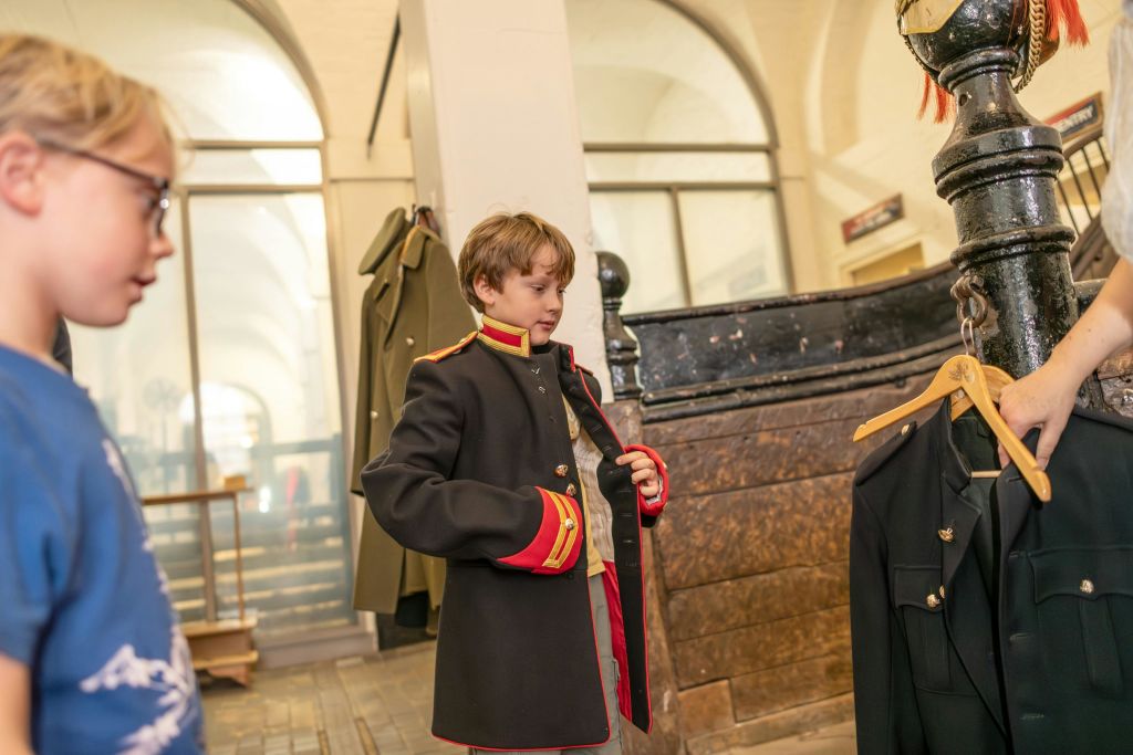 실내에서 역사적인 밀리터리 스타일의 코트를 입어보는 아이와 박물관 같은 분위기에서 관찰하는 또 다른 아이.