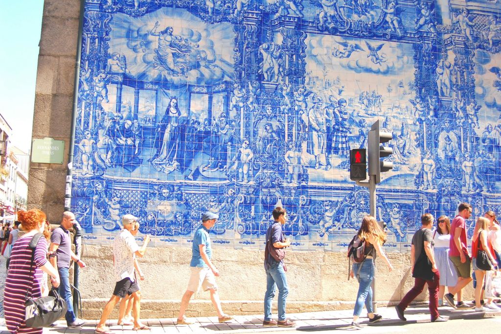 빨간색 보행자 신호등이 있는 커다란 청백색 벽화 벽을 지나 네 사람이 걸어가고 있습니다.