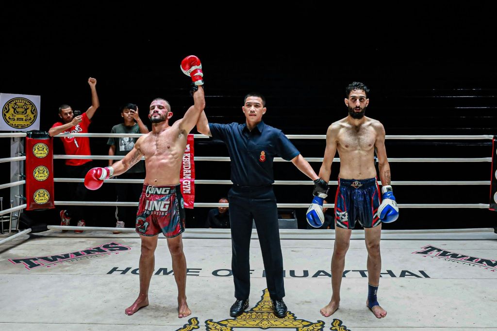 무에타이 선수 2명과 심판 1명이 링에 앉았습니다. 심판이 왼쪽에서 승리한 선수의 손을 들어 올립니다.