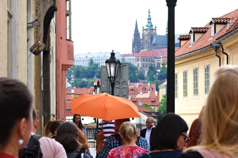 사람들은 유서 깊은 건물과 성을 배경으로 주황색 우산 아래 좁은 거리를 걷고 있습니다.