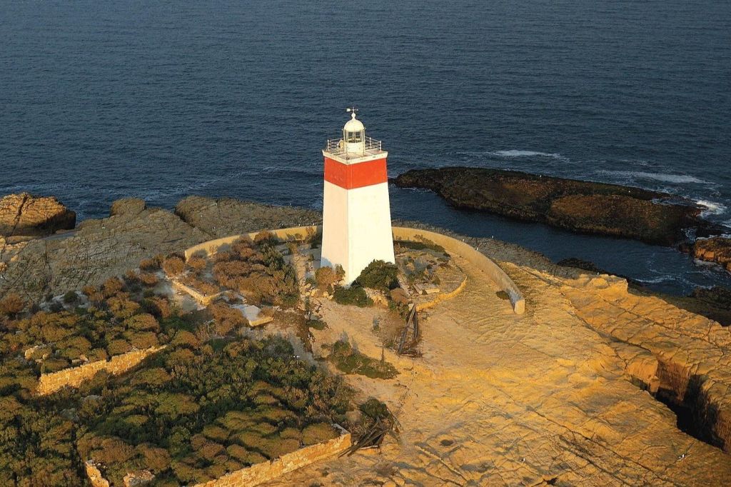 바다 옆에 둘러싸인 초목이 있는 바위 해안 지역의 흰색과 빨간색 등대의 공중 전망.