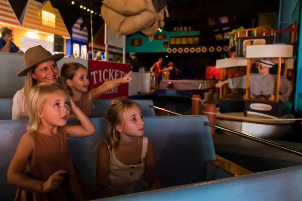 세 명의 어린 소녀와 어른이 있는 가족이 승마 보트에 앉아 밝고 화려한 건물이 있는 놀이공원의 한 장면을 신나게 바라보고 있습니다.