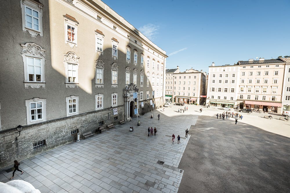맑고 푸른 하늘 아래 사람들이 산책하고 소규모 그룹이 모인 유서 깊은 건물 앞의 햇살 가득한 광장.