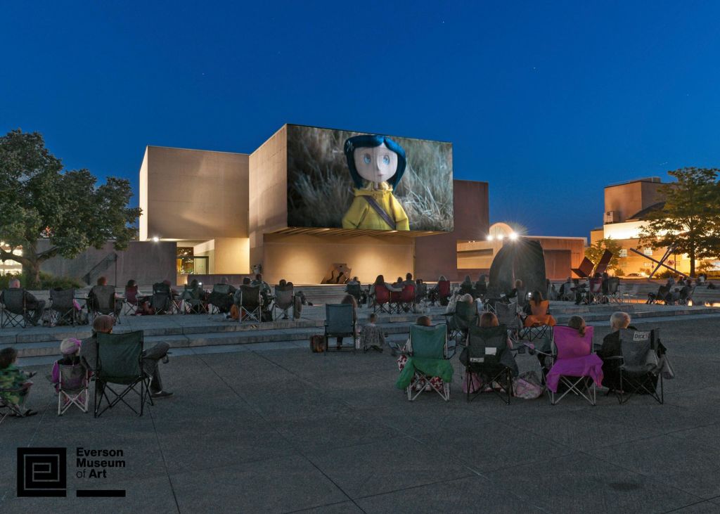 사람들은 야외 의자에 앉아 밤에 현대적인 콘크리트 건물에 부착된 대형 스크린에서 애니메이션 영화를 보고 있습니다.