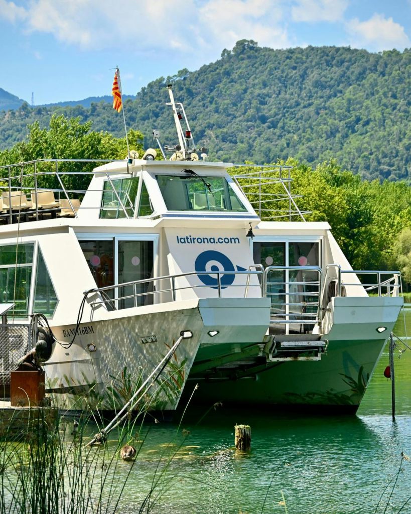 숲이 우거진 언덕을 배경으로 "latirona.com" 이라는 글자가 있는 하얀 쌍동선이 울창하고 푸른 단풍 근처에 정박해 있습니다.