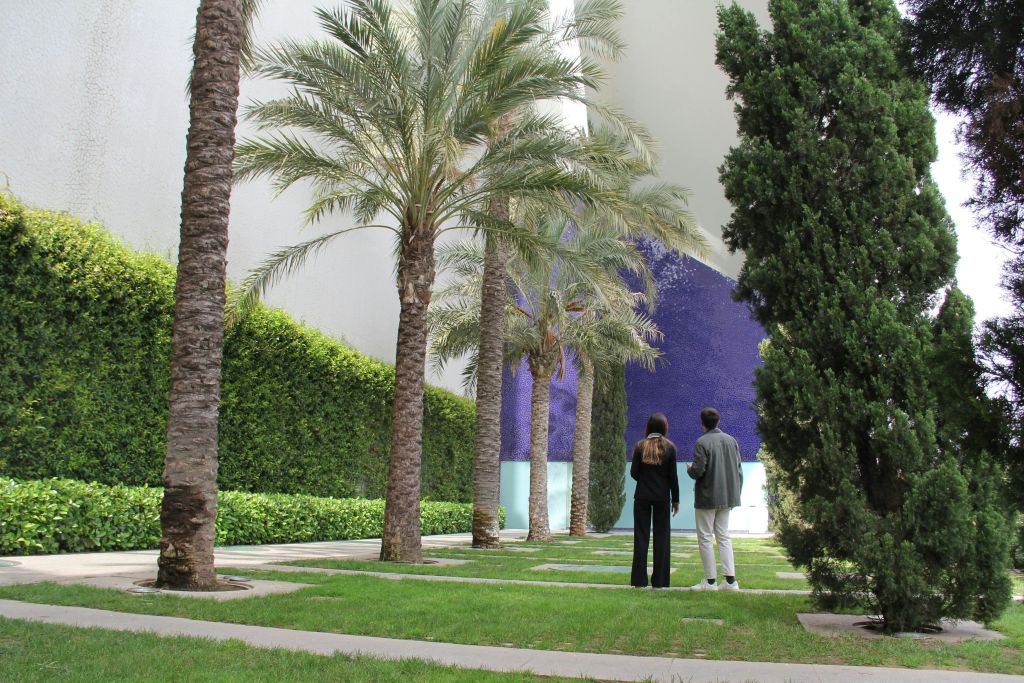 야자수와 커다란 보라색 벽을 배경으로 두 사람이 정원에 서 있습니다.