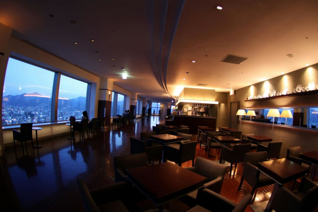 탁자와 의자가 있는 어두운 라운지, 도시 풍경을 보여주는 커다란 창문, 머리 위 조명이 있는 바 공간.