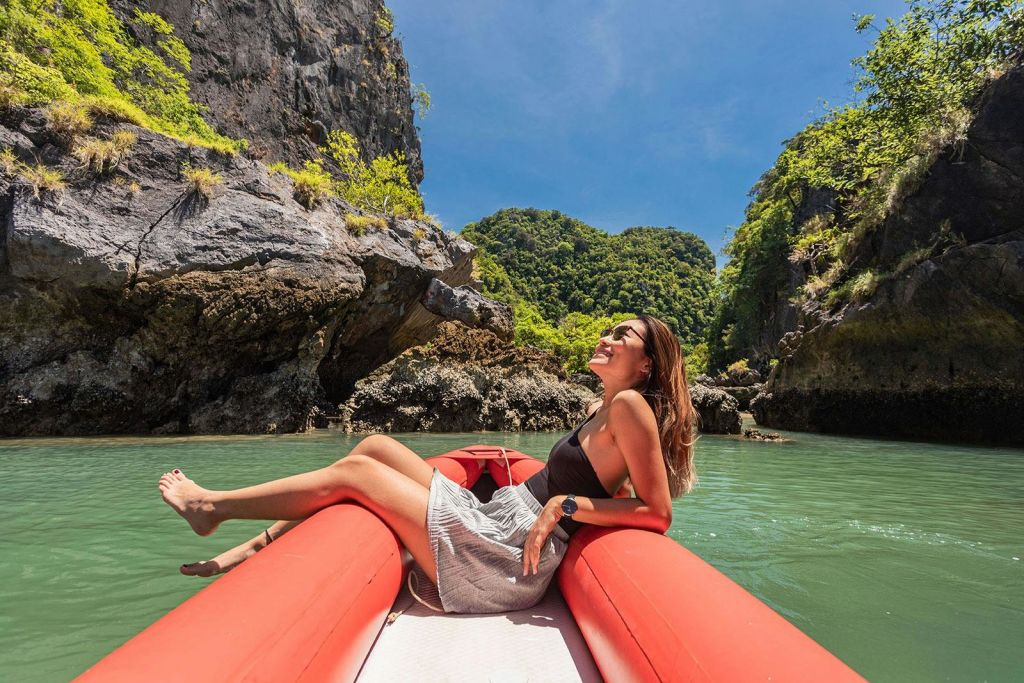 맑고 푸른 하늘 아래 바위 절벽과 푸른 초목이 우거진 경치 좋은 강에서 빨간색 팽창식 보트를 타고 휴식을 취하는 여성.