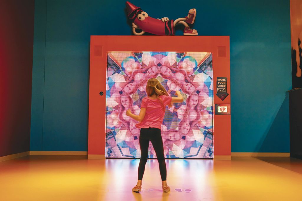 분홍색 옷을 입은 어린이가 크레용 마스코트 아래 파란색 벽을 배경으로 화려한 디지털 디스플레이와 상호 작용합니다.