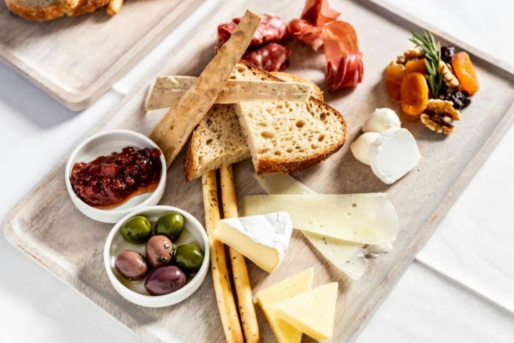 가벼운 나무 표면에 빵 슬라이스, 치즈, 경화 고기, 올리브, 잼, 말린 과일이 있는 샤퀴트리 보드.