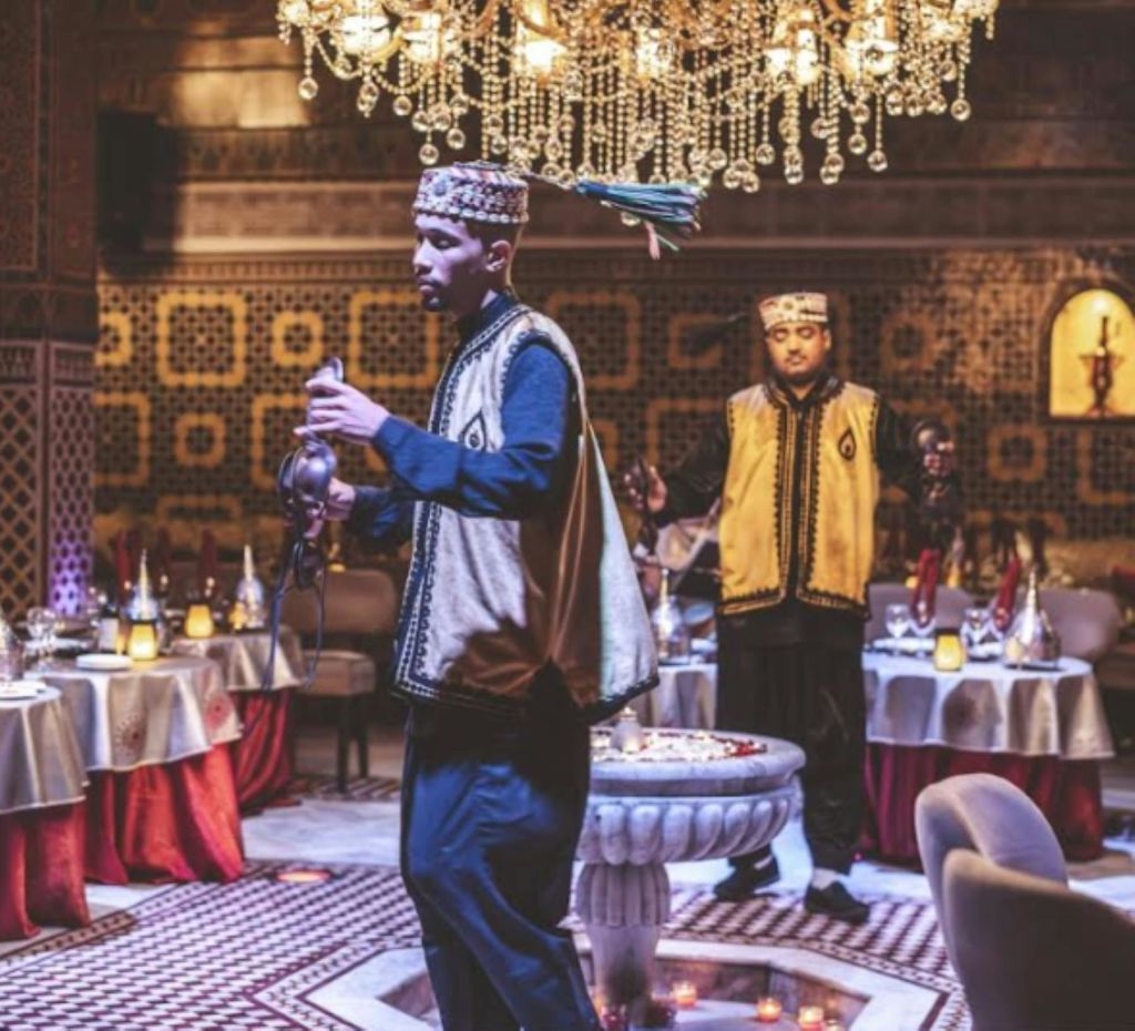 전통 복장을 한 두 남성이 샹들리에와 무늬가 있는 벽이 있는 화려한 레스토랑에서 차를 제공합니다.