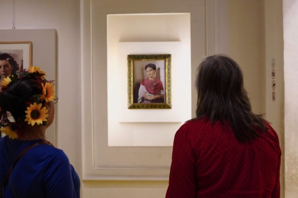 벽에 불이 켜진 틈새에 빨간색과 흰색 드레스를 입은 여성의 액자 그림을 마주한 두 사람이 서 있습니다.