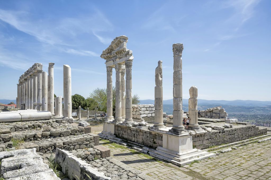 맑고 푸른 하늘 아래 부분적으로 손상되지 않은 높은 석조 기둥이 있는 고대 그리스 유적지. 유적을 배경으로 사람들이 거닐고 있습니다.