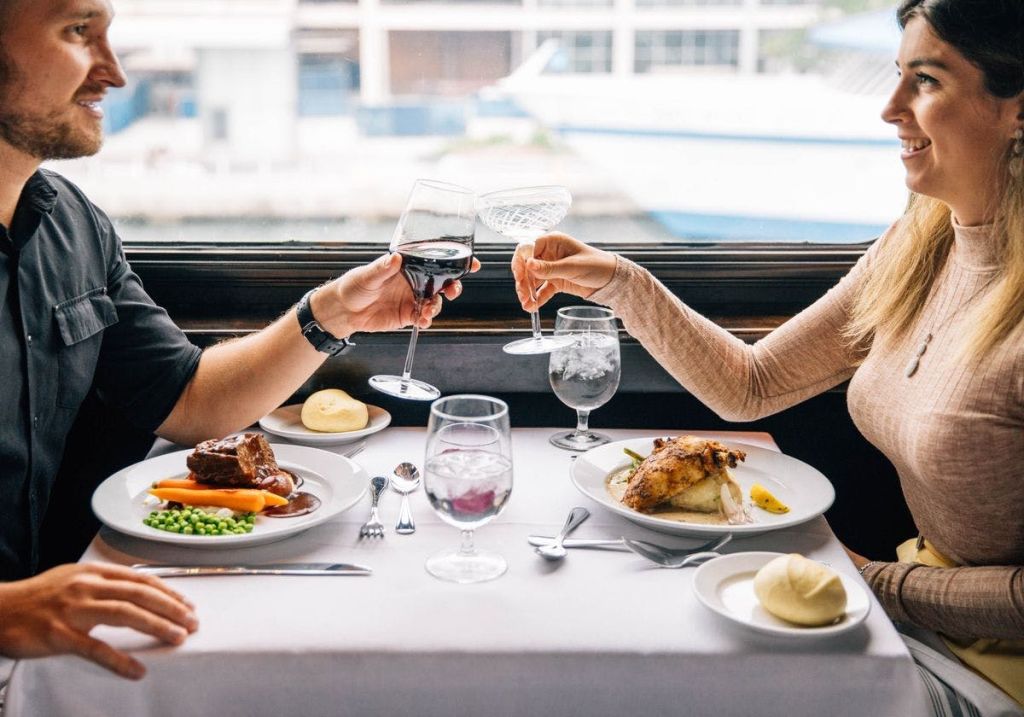 구운 닭고기, 물, 롤 등 음식이 담긴 식탁에서 두 사람이 잔을 움켜쥐고 있습니다. 그들은 웃고 있습니다.