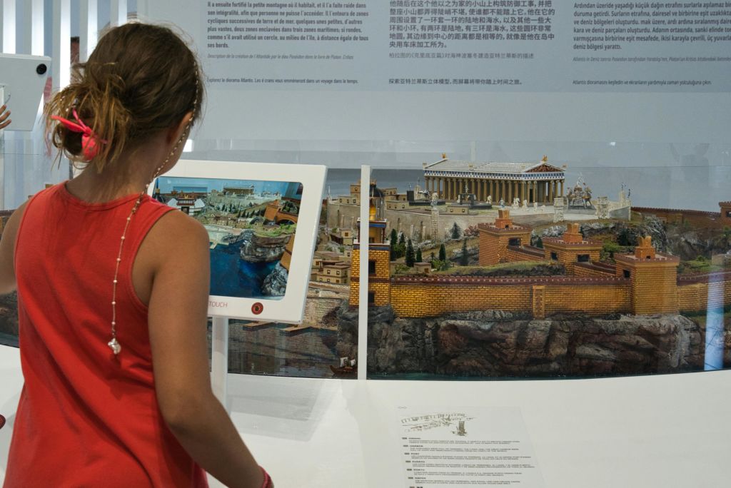 빨간색 민소매 셔츠를 입은 사람이 상세한 역사적 모델을 보여주는 터치스크린 디스플레이와 상호 작용합니다.