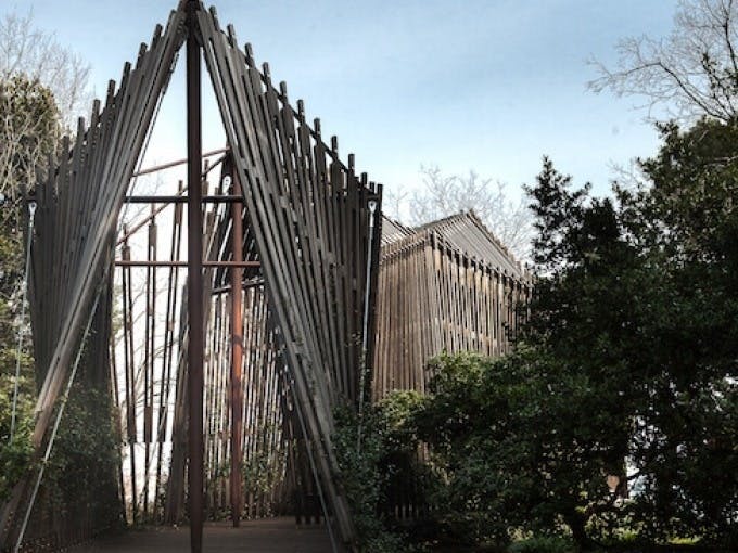 맑은 하늘 아래, 나무로 둘러싸인 삼각형 패턴의 수직 기둥이 있는 목조 구조물.