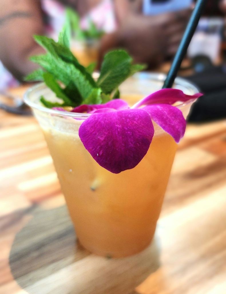 보라색 꽃과 신선한 민트 잎으로 장식된 투명한 컵에 담긴 열대 음료로 빨대가 삽입되어 있습니다.