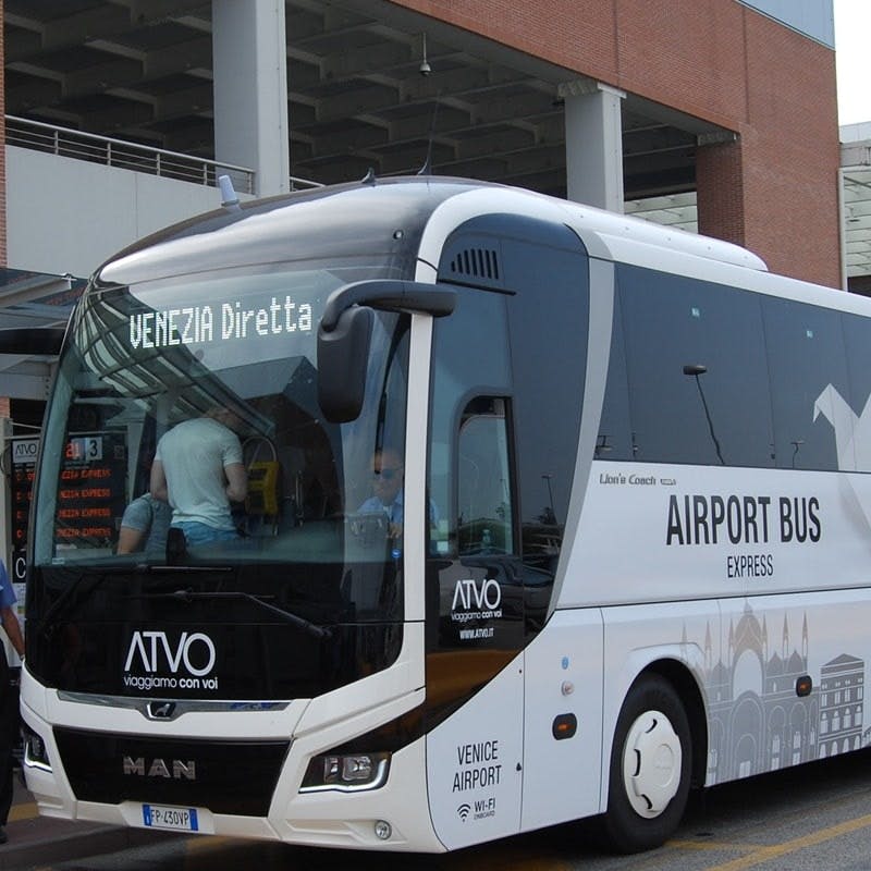 "베네치아 디레타" 와 "공항 버스 익스프레스" 로 표시된 흰색 공항 버스가 건물 외부에 주차되어 있으며 승객이 탑승하고 있습니다.