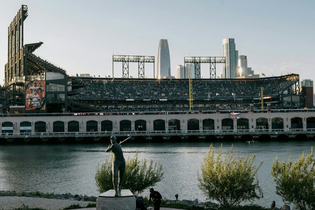 전경에 동상이 있고 도시 고층 건물이 배경으로 보이는 수변 야구 경기장입니다.