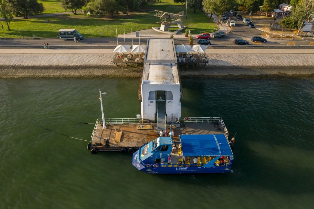 사람들이 탑승하는 부두에 배가 정박했습니다. 부두는 주차된 차량과 인근 나무가 있는 강변 공원에서 뻗어 있습니다.