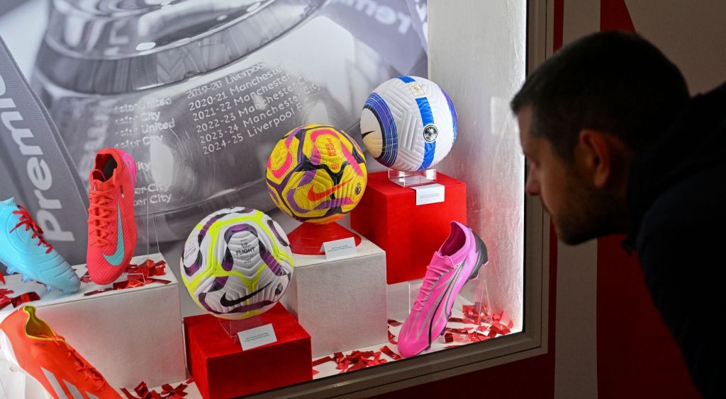 빨간색 받침대에 축구공 3개와 분홍색 축구화. 한 남성이 전시물을 보고 있습니다. 배경 벽에 텍스트.