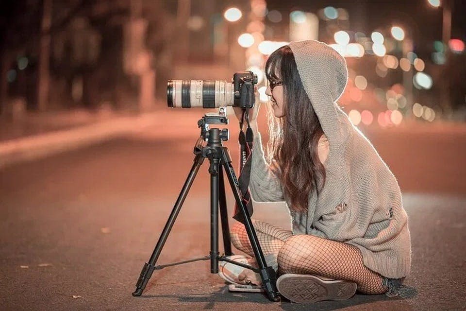 후드티와 망사 스타킹을 입은 사람이 밤길에서 삼각대의 카메라를 들여다보며 무릎을 꿇고 있습니다.