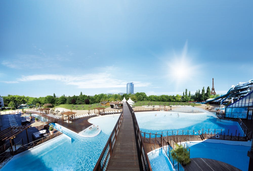 수영장이 있는 대형 야외 워터파크, 나무 산책로, 주변 녹지. 밝은 하늘 아래 배경의 고층건물.