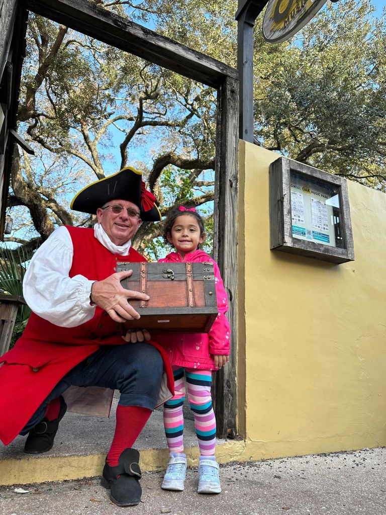 나무 상자를 들고 있는 역사적인 의상을 입은 한 남자가 노란 벽과 나무 출입구 근처에서 어린 소녀 옆에 서 있습니다.