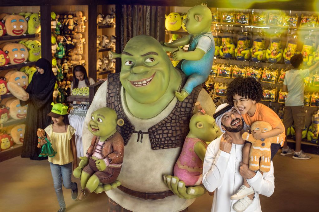 사람들은 슈렉을 테마로 한 상품으로 가득한 밝은 기프트숍에서 슈렉과 그의 가족의 동상과 함께 포즈를 취하고 있습니다.