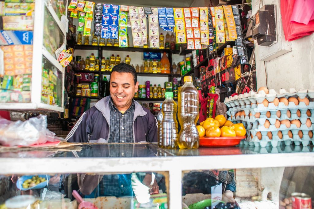 다양한 포장재와 채소, 병이 가득한 작은 가게의 카운터 뒤에 한 남성이 만족스러운 미소를 짓고 있습니다.