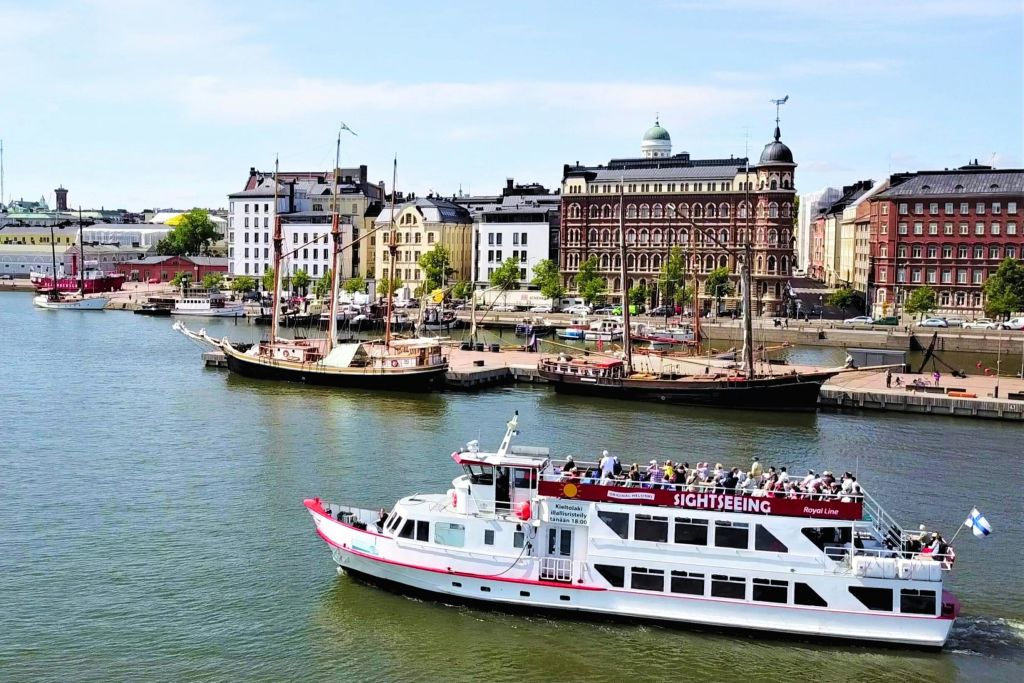유서 깊은 건물과 여러 척의 정박한 항해선을 배경으로 강에서 승객이 항해하는 흰색 관광 보트가 있습니다.