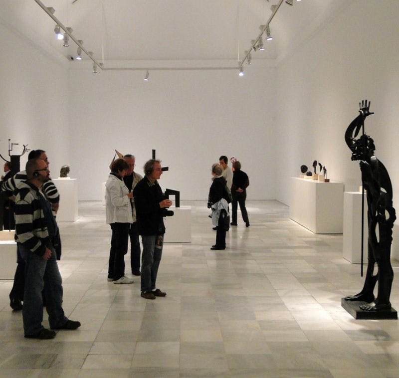 현대 미술관에서 조각품을 감상하는 사람들. 바닥은 타일로 마감되어 있으며, 미술품은 받침대와 선반에 전시되어 있습니다.