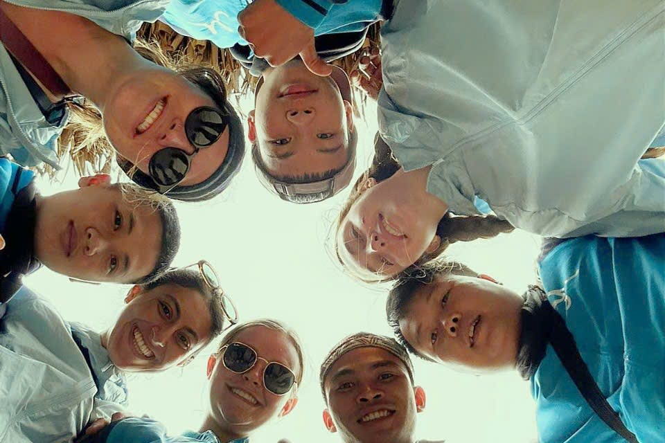 선글라스와 파란색 재킷을 입은 7명의 사람들이 동그랗게 모여 카메라를 내려다보며 미소 짓고 있습니다.