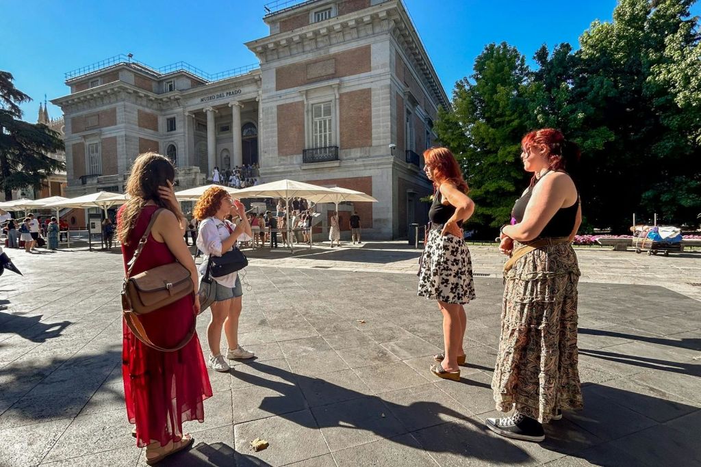 벽돌 건물과 나무 근처의 햇살이 내리쬐는 광장에 서 있는 두 여성과 다른 사람들, 우산 아래 앉은 사람들.