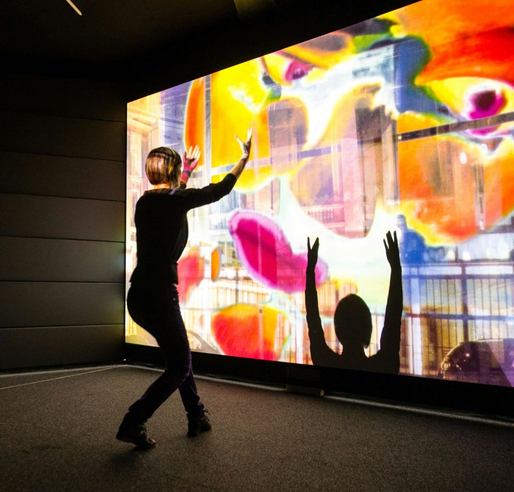 벽에 커다랗고 화려한 디지털 프로젝션과 상호 작용하며 손을 들어 그림자를 드리우는 사람.