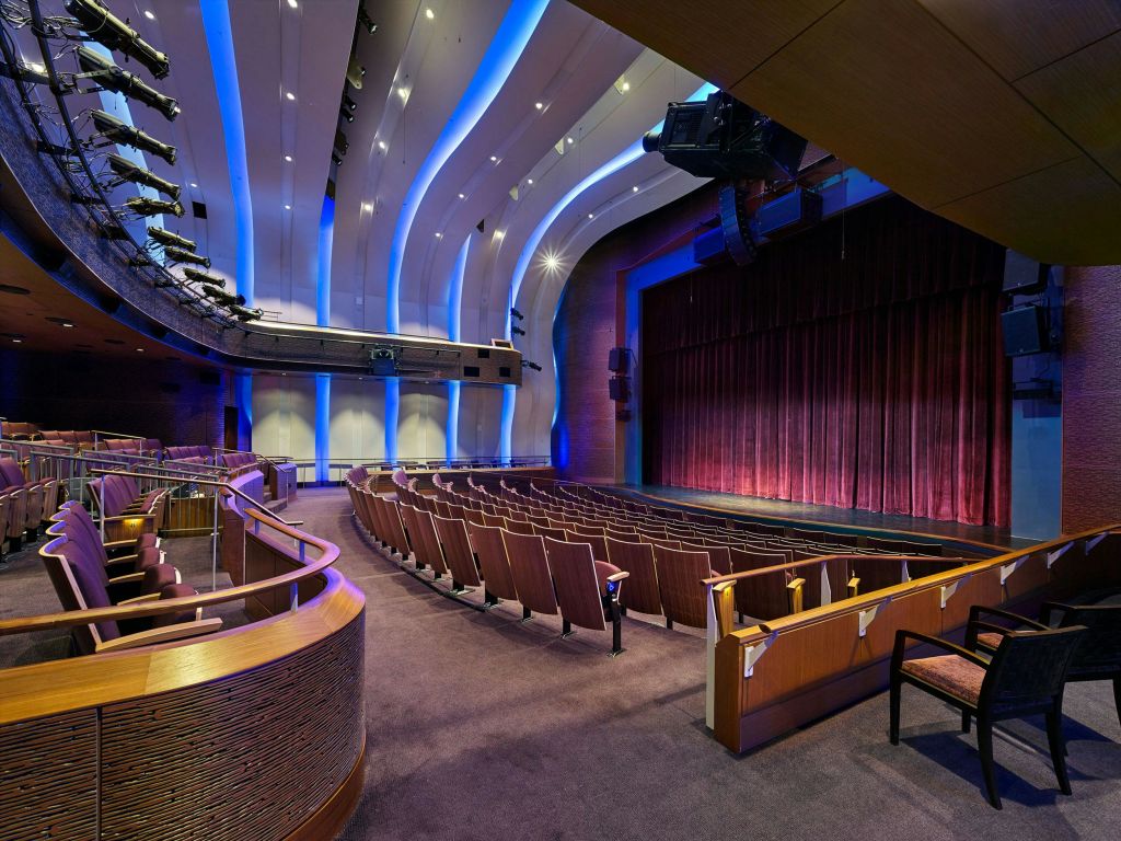 곡선형 좌석이 줄지어 있는 빈 극장 강당, 빨간색 커튼이 있는 대형 무대, 파란색 조명 천장 패널.