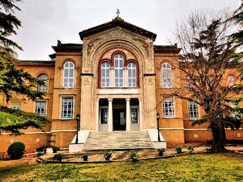 아치형 창문이 있는 크고 오래된 건물, 기둥이 있는 웅장한 입구, 맨 나무와 녹색 잔디밭으로 둘러싸인 돌 계단.