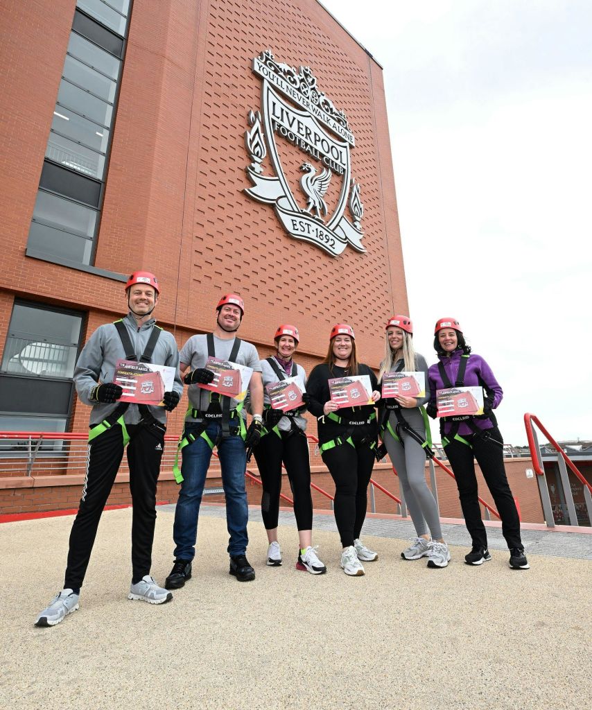 헬멧과 하네스를 쓴 6명이 커다란 리버풀 축구 클럽 문장이 있는 건물 앞에서 수료증을 들고 있습니다.