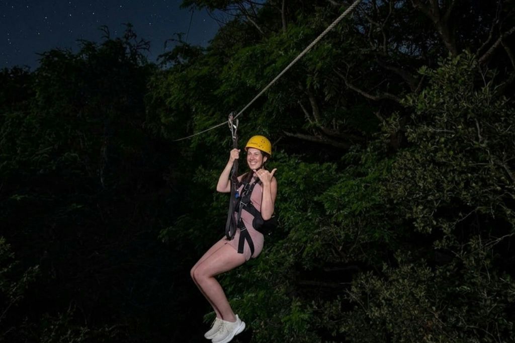 헬멧과 하네스를 착용한 사람이 저녁에 숲을 가로질러 집라인을 하고 있습니다.