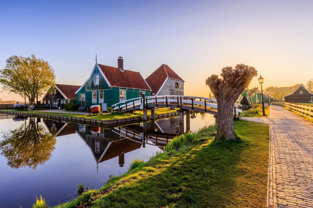 붉은 지붕, 운하를 가로지르는 다리, 해가 질 무렵 풀밭에 있는 맨발의 나무가 있는 온실입니다.